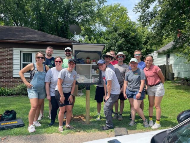 Photograph of a group of ten people posing outdoors around a small, open wooden cabinet filled with books, set on a grassy lawn near residential houses. The scene suggests a community or volunteer activity involving the installation or maintenance of a little free library for public book sharing.