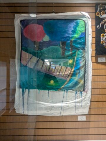 Photograph of a framed abstract painting featuring a landscape with trees, hills, and a bridge, using bold colors like blue, green, and red. The painting has a dripping paint effect at the bottom and is displayed on a wooden slat wall with a small label beneath it.