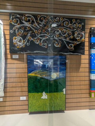 Photograph of two paintings displayed on a wooden slat wall, featuring a stylized tree with swirling branches and circular orange accents above a landscape with a moonlit sky, green hills, and a small white figure. Both artworks use bold colors and abstract elements to evoke nature and night themes, with visible labels beneath each piece.