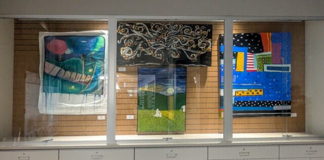 Photograph of a display case featuring three colorful abstract paintings mounted on a wooden panel background. Paintings include a blue and green piece with geometric shapes on the left, a central painting with swirling gold patterns above a moonlit landscape, and a vibrant blue and multicolored geometric composition on the right.