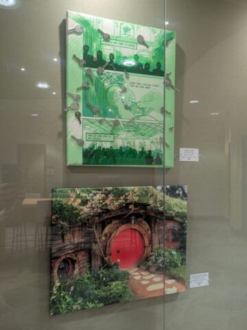 Photograph of two framed artworks displayed on a wall behind glass. Top piece is a green-toned comic panel with multiple keys attached, featuring dialogue and shadowy figures, while bottom piece is a detailed, colorful illustration of a round red door set in a lush, natural environment.