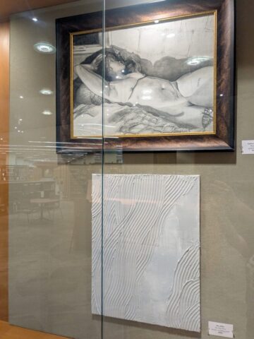 Photograph of two framed artworks displayed on a wall behind glass.