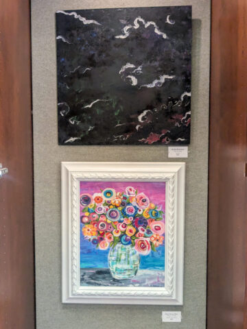 Photograph of two framed paintings displayed vertically on a wall, featuring an abstract dark sky with white cloud-like shapes on top and a colorful bouquet of flowers in a glass vase with a pink and blue background below. The top painting uses dark tones with white highlights, while the bottom painting contrasts with vibrant, circular floral patterns and a decorative white frame.