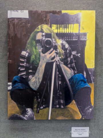 Photograph of a person with green hair sitting on the floor, holding a camera mounted on a tripod, capturing a self-portrait. Background includes a wooden chair and a yellow wall, with the person wearing a plaid shirt, blue jeans, and boots