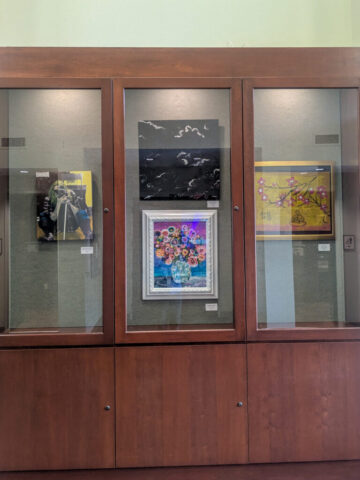 Photograph of a wooden display case with three glass panels showcasing four framed artworks inside. Art pieces include a black and white abstract painting with birds, a colorful floral painting in a white frame, and two other artworks with yellow backgrounds featuring floral and abstract designs.