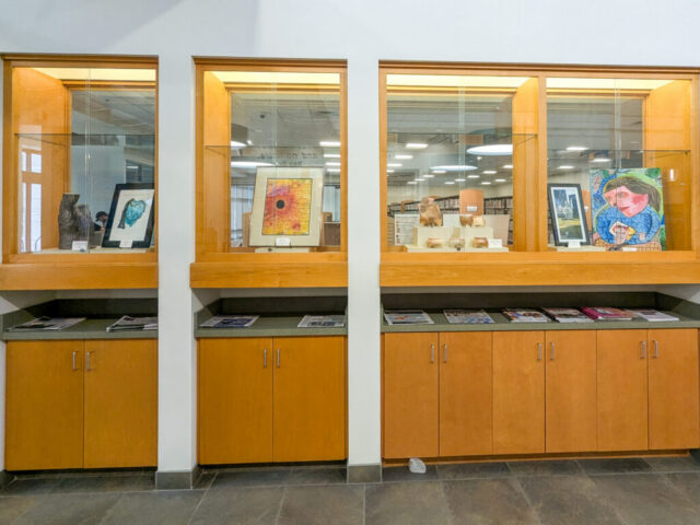 Photograph of a museum or gallery display featuring three wooden and glass cases containing various artworks, including framed paintings and sculptures. Below each case, wooden cabinets hold magazines or brochures, with a tiled floor and white walls providing a clean, organized setting.