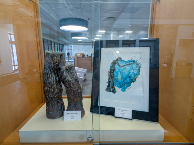 Photograph of a museum display case featuring two art pieces: a textured, dark brown ceramic sculpture resembling tree trunks on the left, and a framed watercolor painting of a bird perched on a large blue heart on the right.