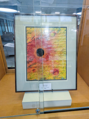 Photograph of a framed abstract painting displayed in a glass case, featuring vibrant red, yellow, and orange colors with circular patterns