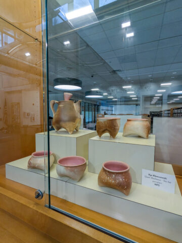 Photograph of a museum display case featuring seven ceramic vessels with rounded bodies and small legs, showcasing ancient pottery craftsmanship. Vessels vary in shape and size with a reddish interior and beige exterior.