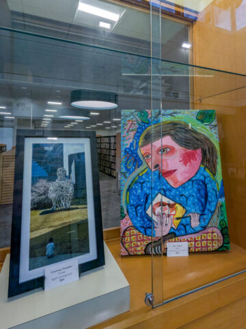 Photograph of an art display case featuring two framed artworks inside a library setting. One artwork is a monochrome wireframe figure sitting outdoors, and the other is a colorful abstract painting of a person.