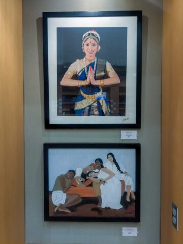 Photograph of two framed artworks displayed on a wall, one above the other. The top artwork depicts a person in traditional Indian dance attire with hands in a namaste gesture, while the bottom artwork shows a group of four people in relaxed poses with earthy tones and simple clothing.