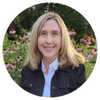 Photograph of a smiling woman with blonde hair wearing a black jacket and white shirt, standing outdoors in front of pink coneflowers and green foliage. The circular frame focuses on her face and upper body, highlighting a natural, serene garden setting.