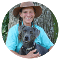Photograph of a smiling person wearing a wide-brimmed hat and blue shirt sitting in front of a tree while holding a gray dog with a collar. The setting appears outdoors with greenery in the background, highlighting a calm and friendly interaction between the person and the dog.