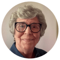 Photograph of an elderly woman with short gray hair and black-framed glasses, smiling gently at the camera. She is wearing a dark blue collared shirt and is positioned against a plain light-colored background.